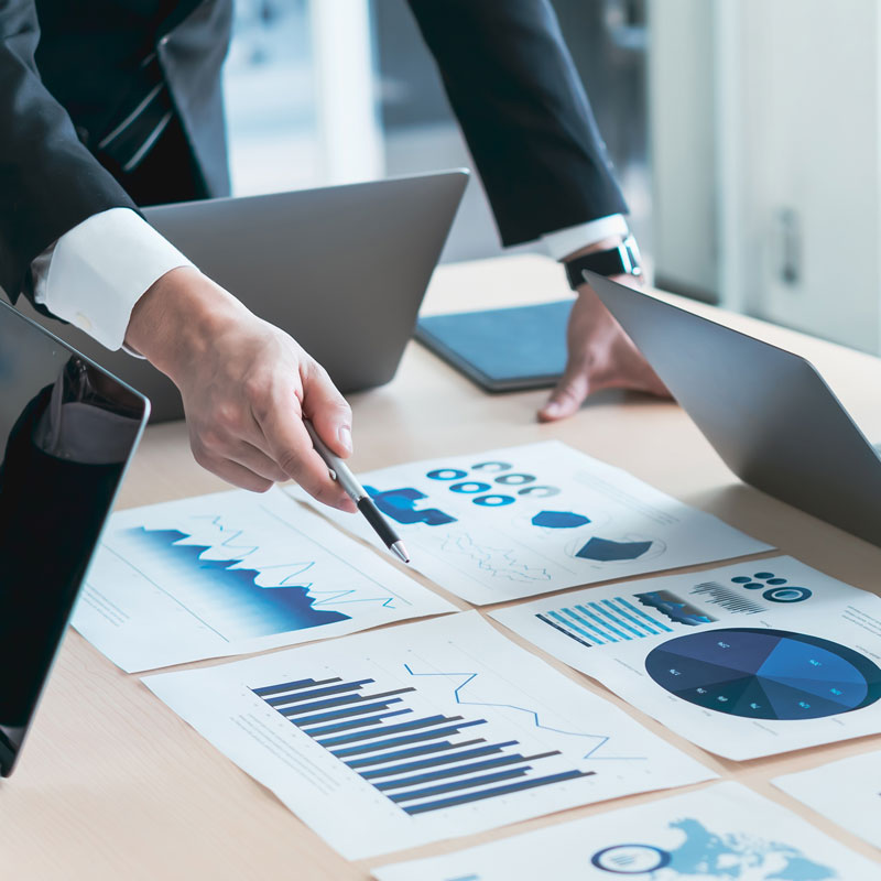 ein Herr im Anzug zeigt mit einem Stift auf Grafiken, die auf einem Tisch liegen